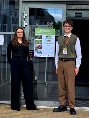 Dwie osoby stoją przed wejściem do budynku, obok plakatu informującego o Konferencji Naukowej