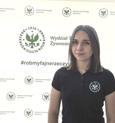 Prezes KNTŻ - Patrycja Chojecka w czarnej koszulce z logo Wydziału Technologii Żywności SGGW, stojąca na tle ścianki wydziałowej.