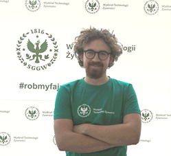 Wiceprezes KNTŻ - Dawid Chróstowski w zielonej koszulce z logo Wydziału Technologii Żywności SGGW, stojący na tle ścianki wydziałowej.